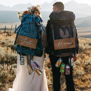 May include: A couple, viewed from behind, wears backpacks and holds wooden signs reading "Just" and "Married". Cans of fizzy drinks hang from the packs. The bride has a floral crown. The scene is set in a natural, outdoor environment.