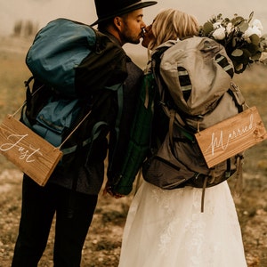 May include: A newly married couple kissing, both wearing backpacks. The person on the left holds a sign saying "Just", the other holds a sign saying "Married". The woman wears a white wedding dress and carries a bouquet of flowers.