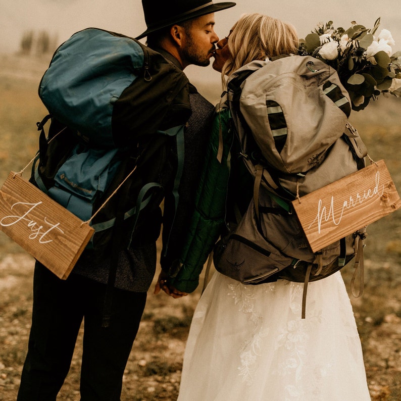 May include: A newly married couple kissing, both wearing backpacks. The person on the left holds a sign saying "Just", the other holds a sign saying "Married". The woman wears a white wedding dress and carries a bouquet of flowers.