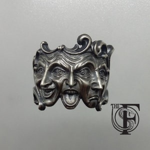 May include: Silver ring with three sculpted faces, each with a different expression. The faces are arranged in a circle, with the top face looking straight ahead, the left face looking to the left, and the right face looking to the right.