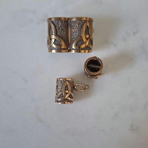 Peut inclure: Un ensemble de trois composants de bijoux en métal de couleur bronze avec une surface texturée et un motif de nœud triquetra en relief. L'ensemble comprend un fermoir rectangulaire large, un fermoir cylindrique et un connecteur tubulaire.
