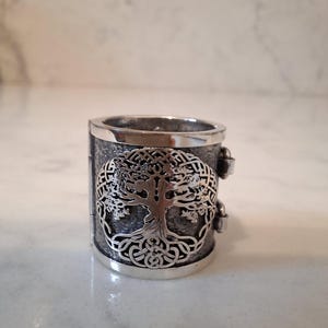 May include: A wide, silver-toned ring featuring a detailed Tree of Life design. The ring has a textured band and decorative clasps on the sides. The Tree of Life is a Celtic symbol, and the ring is likely made of sterling silver.