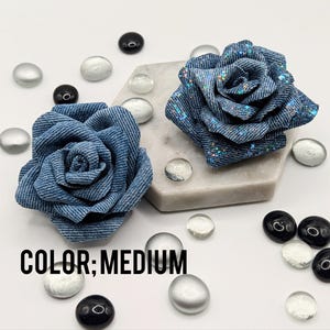 May include: Two denim-style fabric rose hair accessories. One rose sits on a marble hexagon, the other is to the left. Clear, silver, and black decorative glass stones surround the roses. The text "COLOR; MEDIUM" is at the bottom.