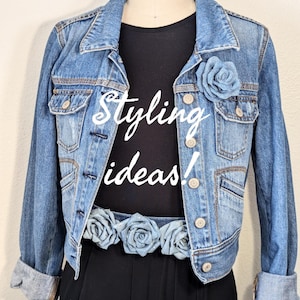 May include: A light blue denim jacket with a black top. The words "Styling ideas!" are written in white script on the top. A blue rose is pinned on the jacket, and a belt with blue roses is around the waist.
