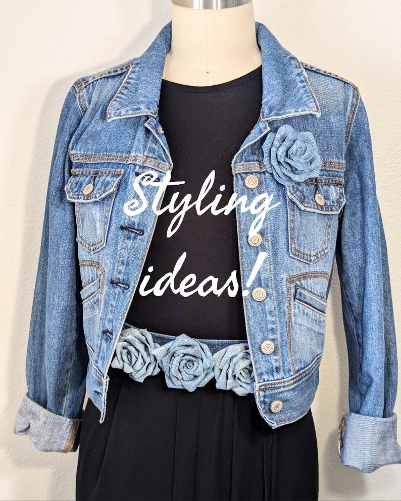 May include: A light blue denim jacket with a black top. The words "Styling ideas!" are written in white script on the top. A blue rose is pinned on the jacket, and a belt with blue roses is around the waist.