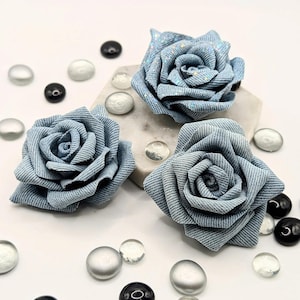 May include: Three light blue fabric roses, resembling denim, with glitter detailing. They are arranged on a white surface with decorative glass beads. The roses have detailed petal construction. The beads are black, clear, and silver.
