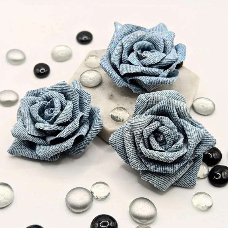 May include: Three light blue fabric roses, resembling denim, with glitter detailing. They are arranged on a white surface with decorative glass beads. The roses have detailed petal construction. The beads are black, clear, and silver.