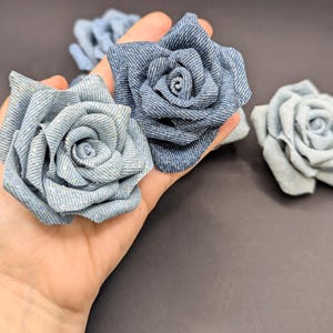May include: Close-up of several denim fabric roses in varying shades of blue. The roses are handmade, with detailed petal construction. The background is a dark grey, providing contrast to the flowers.