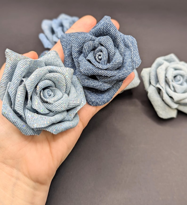 May include: Close-up of several denim fabric roses in varying shades of blue. The roses are handmade, with detailed petal construction. The background is a dark grey, providing contrast to the flowers.