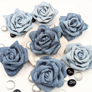 May include: A collection of denim rose flower embellishments in various shades of blue. Some roses have glitter accents. The fabric roses are arranged on a white surface with decorative glass beads.