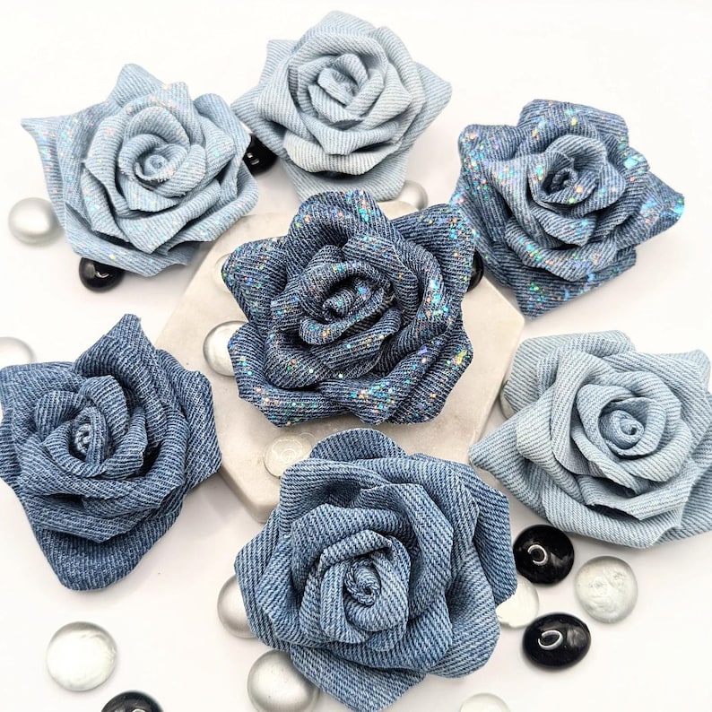 May include: A collection of denim rose flower embellishments in various shades of blue. Some roses have glitter accents. The fabric roses are arranged on a white surface with decorative glass beads.