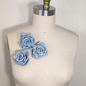 May include: Three light blue fabric roses are pinned to a mannequin. The roses are made of a textured fabric and have a spiral design. The mannequin is a neutral beige colour and has a metal and gold-coloured top.