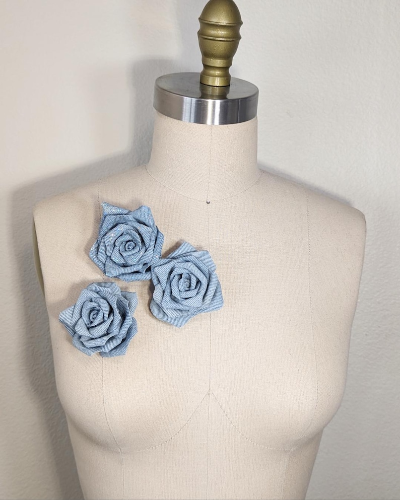 May include: Three light blue fabric roses are pinned to a mannequin. The roses are made of a textured fabric and have a spiral design. The mannequin is a neutral beige colour and has a metal and gold-coloured top.
