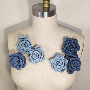 May include: A display of denim rose embellishments in shades of blue, arranged on a mannequin. The roses are made from denim fabric, with some having a sparkly finish. The mannequin is a neutral beige colour.