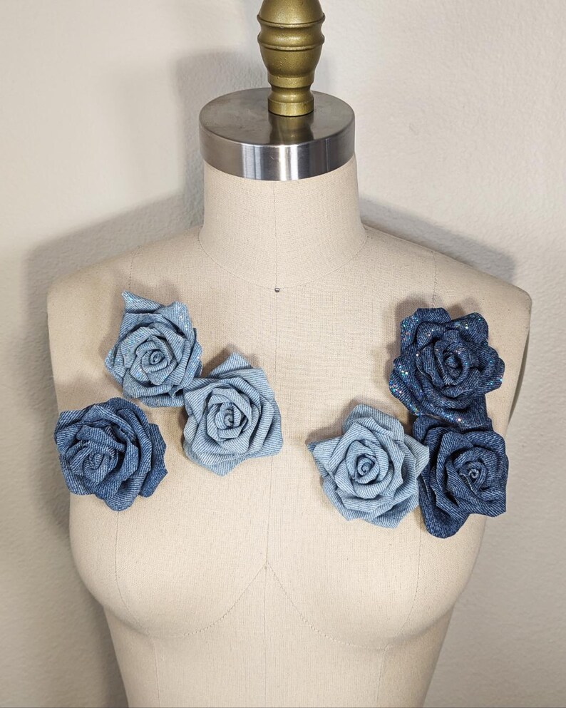 May include: A display of denim rose embellishments in shades of blue, arranged on a mannequin. The roses are made from denim fabric, with some having a sparkly finish. The mannequin is a neutral beige colour.