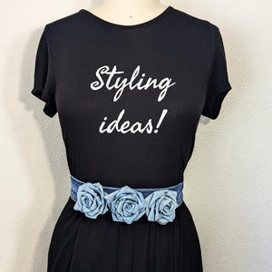 May include: Black t-shirt with the text "Styling ideas!" in white script. A denim belt with three light blue fabric roses is worn around the waist. The outfit is displayed on a mannequin.