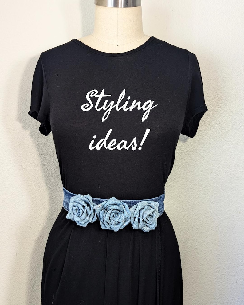 May include: Black t-shirt with the text "Styling ideas!" in white script. A denim belt with three light blue fabric roses is worn around the waist. The outfit is displayed on a mannequin.