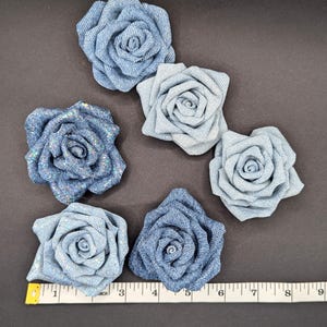 May include: Six denim fabric rose embellishments in varying shades of blue. The roses are arranged on a dark surface with a measuring tape showing inches. The roses range from 5 to 10 cm in diameter.