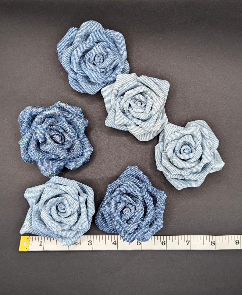 May include: Six denim fabric rose embellishments in varying shades of blue. The roses are arranged on a dark surface with a measuring tape showing inches. The roses range from 5 to 10 cm in diameter.