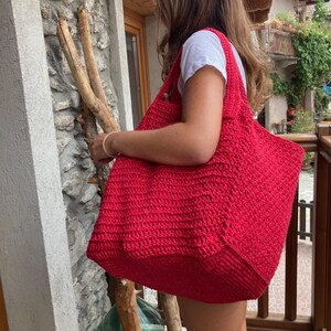 Op de afbeelding: Een grote, felrode gehaakte tote bag met dikke hengsels. De tas wordt vastgehouden door een persoon, maar het gezicht van de persoon is niet zichtbaar.