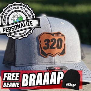 May include: A gray and black trucker hat with a brown leather patch that says "AIELLO 320". The hat is on a concrete surface with a black beanie that says "BRAAAP" in white letters.