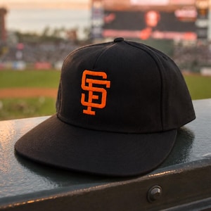 May include: A black baseball cap featuring an embroidered orange SF logo on the front panel. The cap has a structured crown and a flat brim, showcasing a classic athletic design with bold contrast stitching and a clean, minimalist aesthetic.