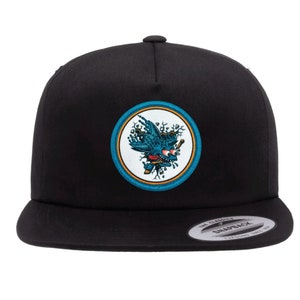 May include: Black and blue embroidered patch on a black snap back hat. The patch features a blue bird with a red eye and a yellow beak.