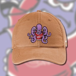 May include: A brown baseball cap with an embroidered purple octopus wearing a red shirt. The octopus has eight tentacles and is in a fighting pose.