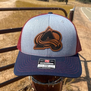 May include: A gray and red baseball cap with a brown leather patch featuring the Colorado Avalanche logo. The cap is a trucker style with a mesh back and a curved brim. The patch is stitched onto the front of the cap. The cap is sitting on a rusty metal post with barbed wire.