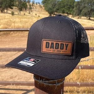 May include: Black and brown trucker hat with a brown leather patch that says "Daddy - Est. 2024".