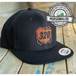 May include: Black and brown baseball cap with a brown leather patch that says "Aiello 320". The cap has a black brim and a white sticker on the underside of the brim that says "The Authentic Snapback".