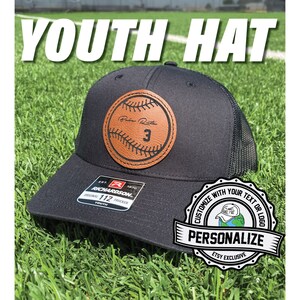 May include: Black youth baseball cap with a brown leather patch featuring a baseball and the text "Rabe Rith 3". The cap is a trucker style with a mesh back. The cap is personalized with the text "CUSTOMIZE WITH YOUR TEXT OR LOGO" and "PERSONALIZE" in a white circle with a black border.
