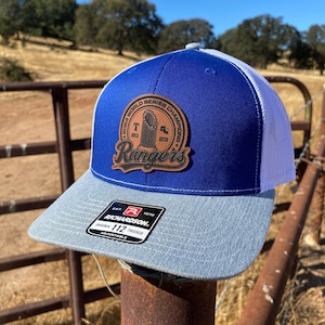 May include: A blue and white trucker hat with a gray brim. The hat features a leather patch with the text "2023 World Series Champions Rangers". The hat is adjustable.