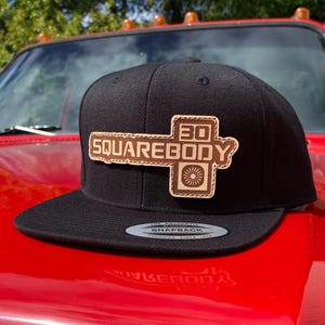 May include: A black snapback cap featuring a tan leather patch on the front. The patch displays the text 30 SQUAREBODY and a circular graphic. A silver and black sticker is visible on the flat brim of the cap, which has a structured crown.