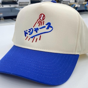 May include: A white baseball cap with a blue brim. The front of the cap features a red and blue embroidered logo with the Japanese text "ドジャース" and a baseball.