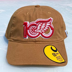 May include: A tan baseball cap featuring an embroidered red and white logo of the number 100 with a winged wheel. A yellow circular sticker on the brim displays the text Force, Wicks Sweat and Dries Fast, and a small white tag is on the side.