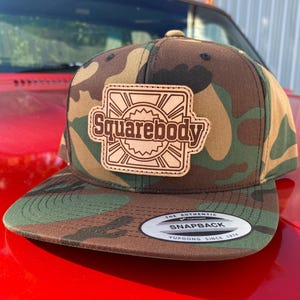 May include: A camouflage print snapback cap featuring a rectangular brown leather patch on the front. The patch displays the text Squarebody centered within a geometric sunburst design. A circular sticker on the flat brim reads The Authentic Yupoong.