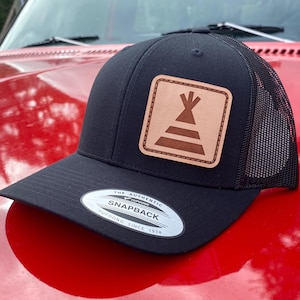 May include: Black trucker hat with a mesh back and a flat brim. The hat features a square, tan leather patch with a brown teepee design. The hat has the text "THE AUTHENTIC SNAPBACK YUPOONG SINCE 1974" on the front.