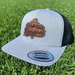 May include: A light blue and black baseball cap with a brown leather patch that says "World Series Champions Texas Rangers". The cap has a black mesh back and a white label that says "The Authentic Snapback Yupoong Since 1974".