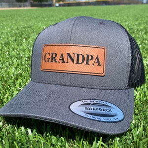 May include: A gray and black baseball cap with a brown leather patch that says "GRANDPA". The cap has a black mesh back and a white sticker that says "THE AUTHENTIC SNAPBACK YUPOONG SINCE 1974".