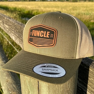May include: A green and beige baseball cap with a brown leather patch that says "FUNCLE 20 24" on the front. The cap has a mesh back and a white sticker that says "THE AUTHENTIC YUPOONG SNAPBACK YUPOONG SINCE 1974".