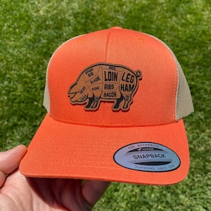May include: An orange trucker hat with a tan mesh back. A brown leather patch on the front features a pig illustration with labeled cuts of meat. The hat has a snapback closure and a "YUPOONG SINCE 1974" tag.