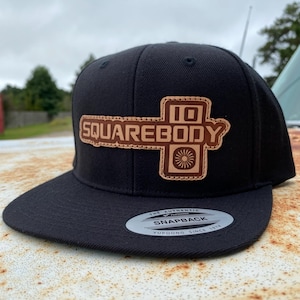May include: Black snapback hat with a flat brim. The hat features a brown leather patch with the words "SQUAREBODY" and "10" in a stylized font. The patch also includes a sunburst design. The hat has a "Snapback" label.