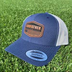 May include: A navy blue and white trucker hat featuring a brown leather-like patch on the front. The patch displays the text GODFATHER EST. 2024 above a stylized American flag graphic. A black and silver sticker on the brim reads THE AUTHENTIC YUPOONG SNAPBACK.