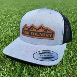 May include: A gray and black trucker hat with a brown leather patch that says "HIGH COUNTRY" with a mountain range design. The hat has a black mesh back and a YUPOONG SNAPBACK label.