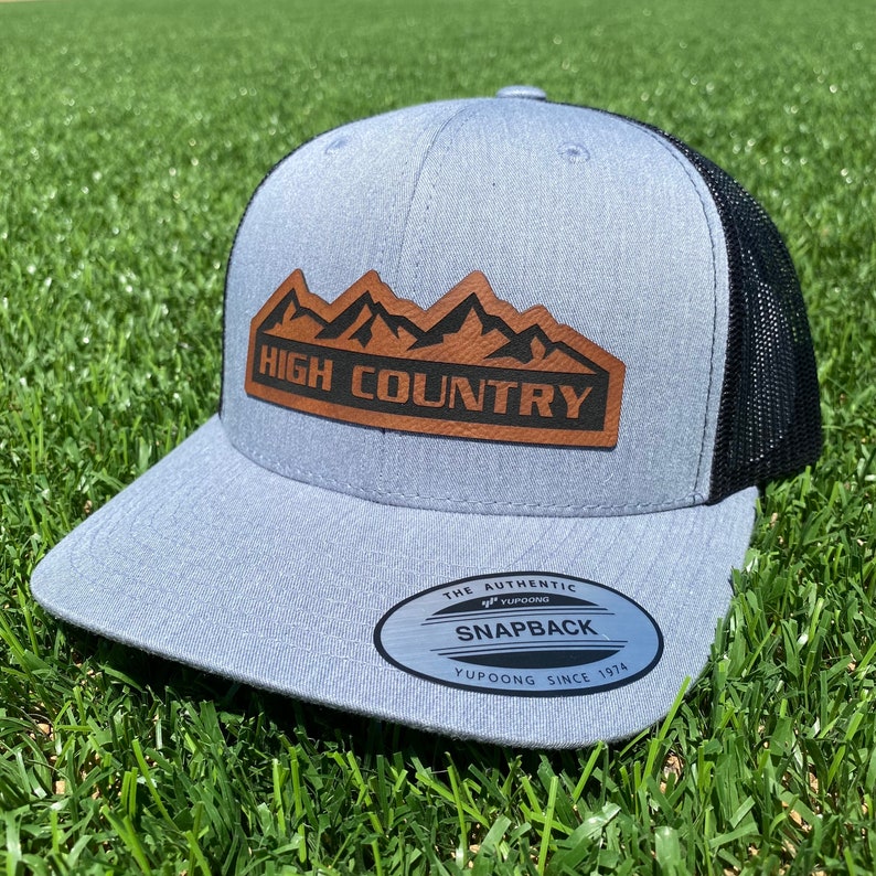 May include: A gray and black trucker hat with a brown leather patch that says "HIGH COUNTRY" with a mountain range design. The hat has a black mesh back and a YUPOONG SNAPBACK label.