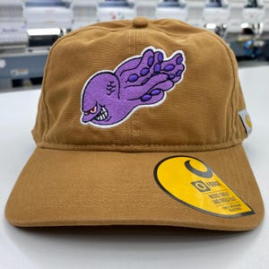 May include: A tan baseball cap featuring an embroidered purple octopus graphic on the front panel. The cap includes a yellow circular sticker on the brim with the text Force, Wicks Sweat and Dries Fast, and a small white tag on the side.