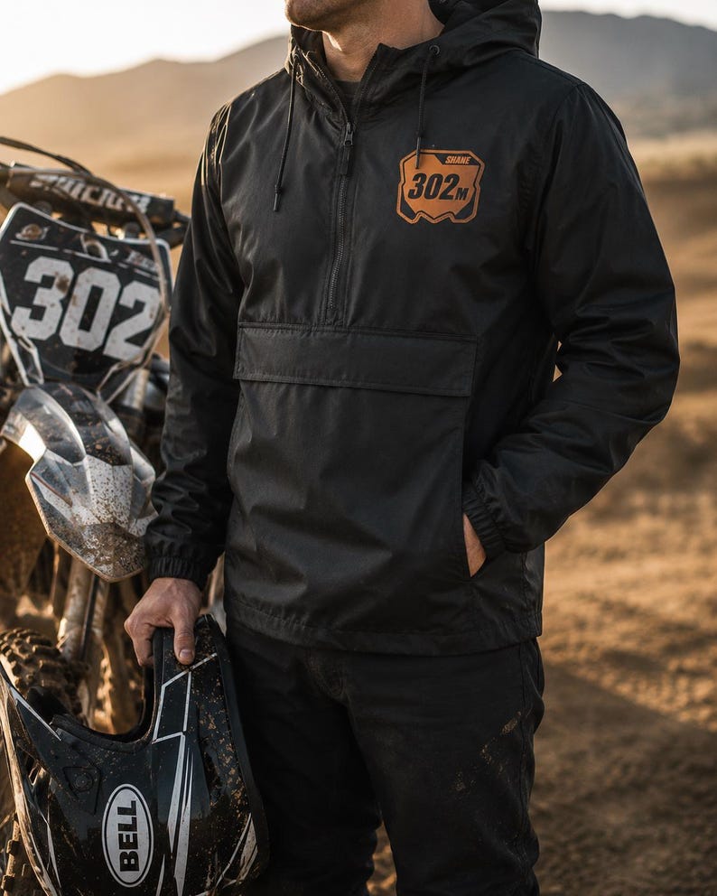 Rider wearing a custom motocross windbreaker with personalized name and number leather patch at an outdoor dirt bike track.