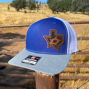 May include: A blue and white trucker hat with a brown leather patch featuring a Texas-shaped outline with a stylized 'D' inside. The hat is adjustable and has a Richardson brand label.