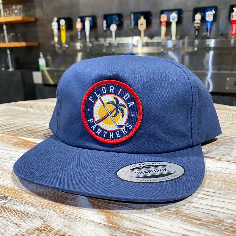 May include: A navy blue baseball cap with a red, yellow, and white Florida Panthers logo. The logo features a palm tree and crossed hockey sticks. The cap has a black and silver sticker that says "The Authentic Snapback Yupoong Since 1974".
